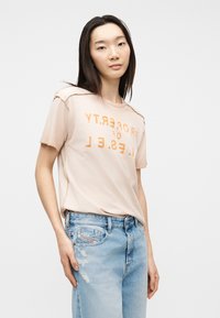 Γυναίκα με ίσια σκούρα μαλλιά, φορώντας μπεζ μπλουζάκι με στάμπα και ανοιχτόχρωμα τζιν με φθορές, στέκεται μπροστά σε απλό λευκό φόντο.