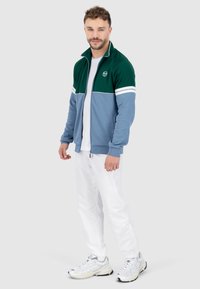 Giacca a track verde e blu, con colletto alto, strisce bianche e logo, abbinata a pantaloni bianchi e sneaker grigie.