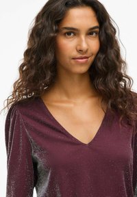 Burgunderfarget topp med V-hals, strikket tekstur og subtil glans. Har strukturert skulderdetalj og langt erme.