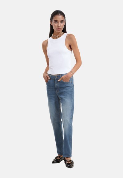 Witte geribde tanktop, lichtblauwe rechtlijnige jeans en zwarte flats, met de handen van het model in de zakken en een gelaagde kettinghanger.
