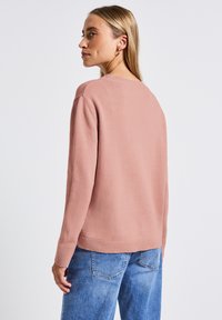 Pull en tricot rose clair avec un col rond, des épaules tombantes et des poignets côtelés ; porté avec un jean bleu, vu de dos.
