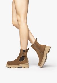 Bottes chelsea en cuire suédé marron avec une fermeture éclair sur le côté, des panneaux élastiques et une semelle en caoutchouc épaisse. Présente un accent décoratif avec une boucle en métal argenté.