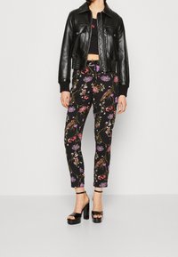 Kobieta ubrana w czarną skórzaną kurtkę, czarne spodnie w kwiatowy wzór, czarny crop top i czarne platformy, stojąca przy białej ścianie.