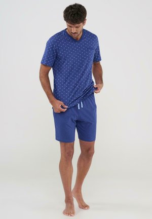 Hombre con camisa azul de manga corta con estampado y cuello en V y pantalones cortos azules con cordón, de pie descalzo, mirando hacia abajo mientras se ajusta la camisa.