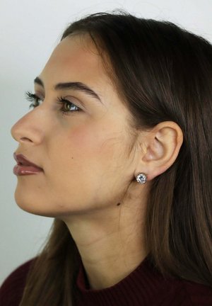 Jonge vrouw met lang bruin haar, gekleed in een donkerrode trui en een enkele ronde diamanten oorbel, kijkt naar links tegen een eenvoudige achtergrond.