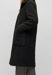 Sort struktureret frakke med en synlig krave, sidelommer og en løs pasform, lavet af et blødt, fleece-agtigt stof. Lange ærmer, knælængde design.