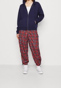 Sudadera con cremallera azul marino con cordones, combinada con pantalones holgados a cuadros rojos que tienen bolsillos, puños elásticos y zapatillas blancas.