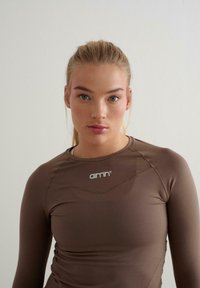 Pruun pika varrukaga sportlik särk, millel on sile tekstuur. Särgil on ümara kaelusega lõige ja rinnal on valge logo.