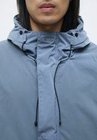 Svetlo modra jakna z visokim ovratnikom, vrvicami in pritisnimi gumbi. Gladek material, na prsnem delu pa je logotip.
