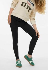 Leggings noirs en tissu extensible, associés à un sweat-beige avec un texte imprimé et des baskets vertes avec un accent blanc.