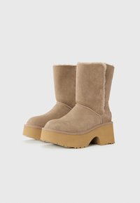 Beige suède enkellaarzen met een dikke, robuuste beige plateauzool. Het interieur is voorzien van een zachte voering, met zichtbare stiksels en subtiele branding.