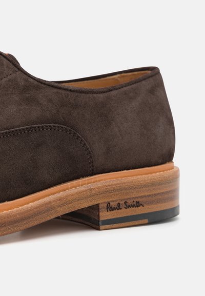 Chaussure en daim marron avec un design arrondi, semelle contrastante couleur tan et talon texturé. Présente le logo 'Paul Smith' sur le côté.