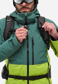 Zielono-żółta wodoodporna kurtka z wzorem w kratę, z przednimi zamkami błyskawicznymi i regulowanymi paskami. W zestawie czarny plecak i rękawice.