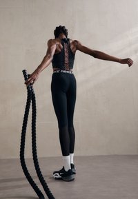 Czarny sportowy top na ramiączkach z koronkowym detalem na plecach oraz czarne legginsy z białym pasem, połączone z czarnymi butami sportowymi i liną do ćwiczeń.