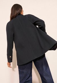 Melnas, rūpīgi šūtas mētelis ar drapētu dizainu, ar garām piedurknēm un vienu pogu aprocē. Gluda tekstūra un strukturēta piegriezne.