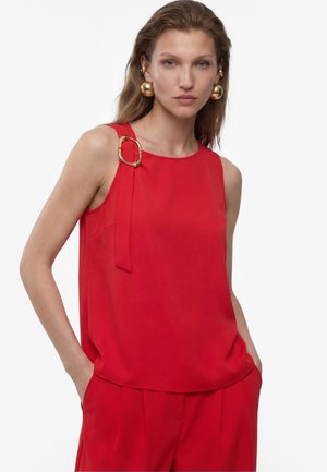Femme portant un haut rouge sans manches avec un détail de boucle dorée à l'épaule, pantalon rouge assorti et grandes boucles d'oreilles dorées, mains dans les poches.