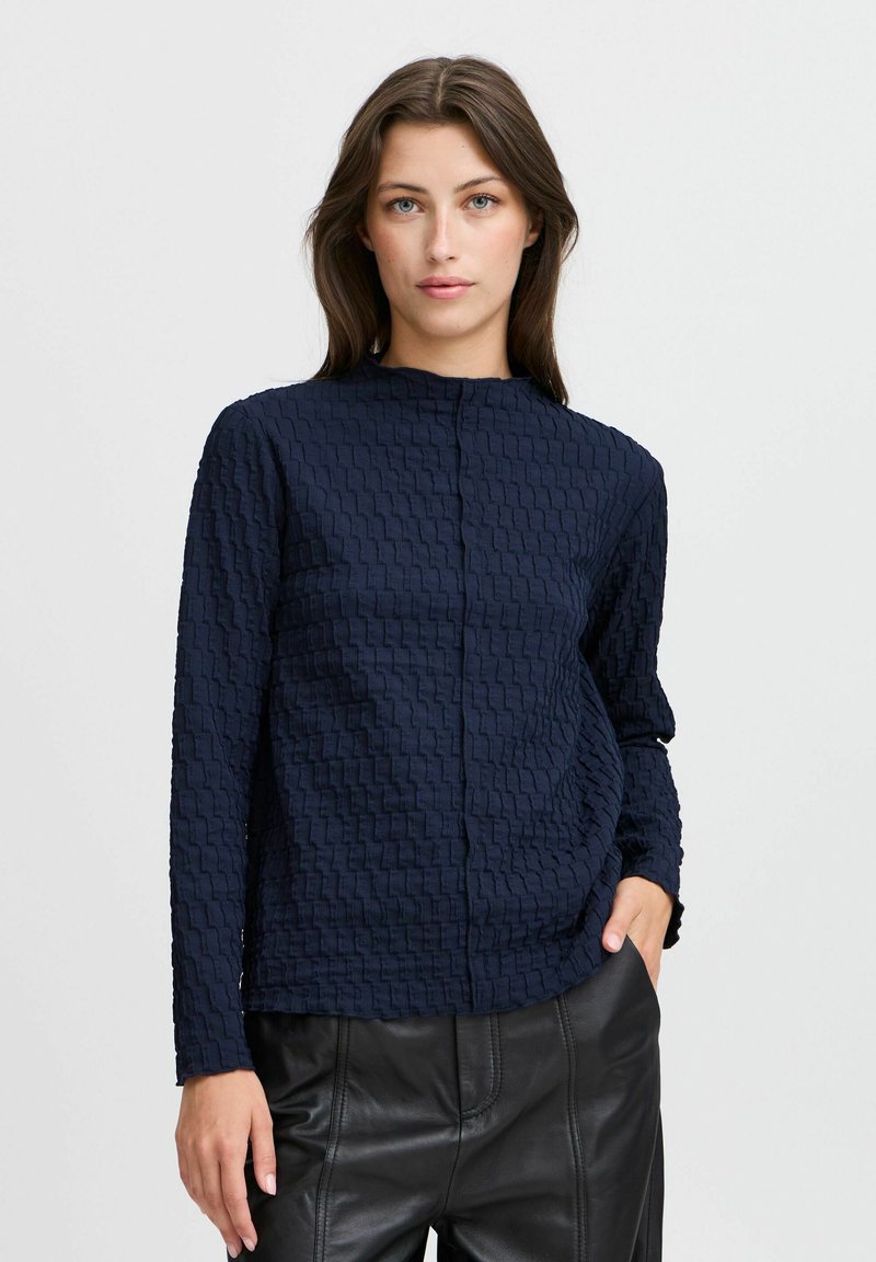 Haut bleu marine texturé à manches longues avec un col haut et des motifs géométriques en relief, associé à un pantalon en cuir noir.