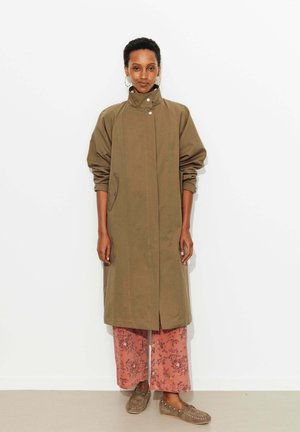 Femme portant un long manteau vert olive, un pantalon large à fleurs roses, et des mocassins, debout devant un fond blanc uni.