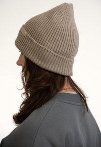 Beige ribbed knit beanie with a pointed top and a folded cuff, worn at a slightly tilted angle. Soft texture and subtle branding on the cuff.