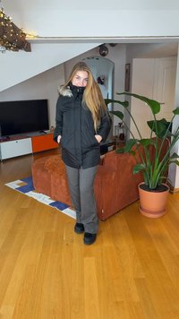 Veste isolante noire avec capuche doublée de fourrure, fermeture éclair à l'avant et deux poches latérales. Portée avec un pantalon gris et des chaussures noires. Environnement intérieur.