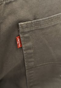 Κοντινή λήψη τσέπης από χακί ύφασμα με εμφανές κόκκινο ετικέτα Levi's και λεπτομερή ραφή στην άκρη της τσέπης.