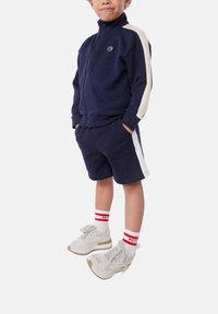 Marineblaue Trainingsanzug mit cremefarbenen Seitenstreifen; Zip-Jacke und passende Shorts. Graue Turnschuhe mit rot-weißen gestreiften Socken.