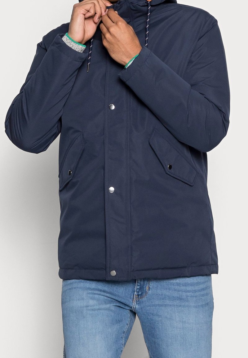 Mörkblå jacka med huva, tryckknappar, två sidofickor och dragsnören. Tillverkad av ett slätt, hållbart tyg med en vadderad design.