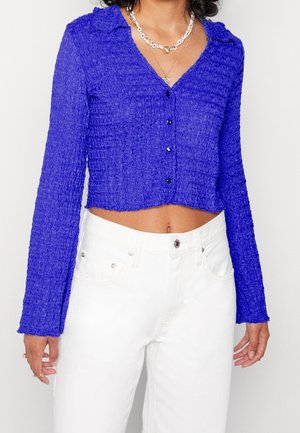Kvinde iført en struktureret kongeblå langærmet knaplukket cropped top med hvide højtaljede jeans og lagdelte delikate halskæder.