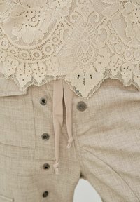 Haut en dentelle beige avec des motifs floraux complexes, superposé à un pantalon en tissu beige clair équipé de boutons et d'une fermeture à cordon.