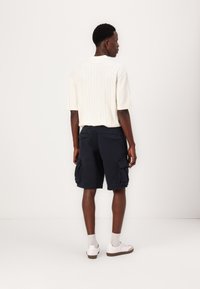 Haut en tricot à rayures blanches à manches courtes associé à un short cargo bleu marine foncé avec de nombreuses poches. Des baskets blanches à semelle basse complètent la tenue.