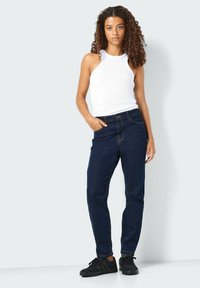 Temno modri jeans z ohlapnim krojem, v kombinaciji z belim rebrastim topom brez rokavov. Črne superge dopolnjujejo obleko in izpostavljajo sproščen slog.