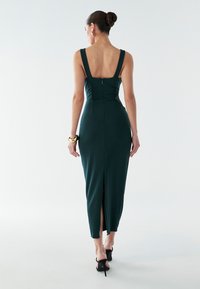 Ciemnozielona dopasowana sukienka z szerokimi ramiączkami, niskim dekoltem na plecach i zamkiem z tyłu; posiada wysoki rozcięcie przy krawędzi. Gładka tekstura.
