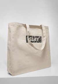 Sac fourre-tout en toile beige avec deux poignées et un patch rectangulaire noir avec un motif paisley blanc et le mot « SLAY » en lettres grasses.