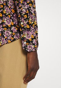 Main à la peau foncée reposant sur le côté, portant une chemise noire avec un motif floral violet et orange et un pantalon beige sur fond blanc.