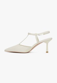 Kremowe skórzane szpilki typu slingback z wąskim obcasem i projektem T-strap. Gładka faktura z minimalną ilością detali oraz jasne drewniane akcenty na obcasie.