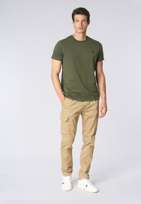 T-shirt verde oliva con maniche corte e un logo sul petto, abbinato a pantaloni cargo beige con più tasche, indossato con sneakers bianche.
