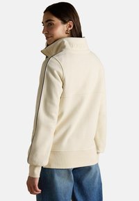 Kremowy polarowy sweter z wysokim kołnierzem, subtelnym wytłoczeniem "JOULES" oraz ciemnym obszyciem po bokach; posiada ribbowe mankiety i dół.