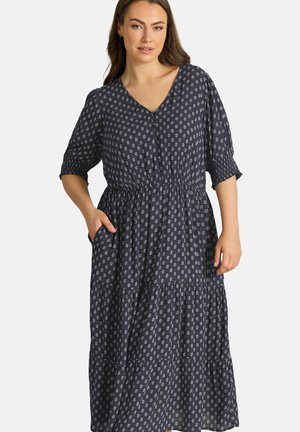 Frau trägt ein dunkelblaues Midikleid mit weißen Blumenmustern, V-Ausschnitt, Dreiviertelärmeln, elastischer Taille und Händen in den Taschen.