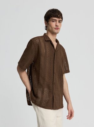 Giovane uomo che indossa una camicia marrone a maniche corte a trama aperta, larga, con bottoni, e pantaloni chiari su uno sfondo semplice.