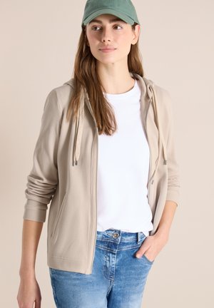 Jeune femme portant une casquette verte, un sweat à capuche zippé beige, un t-shirt blanc et un jean bleu, debout avec une main dans la poche devant un fond beige.