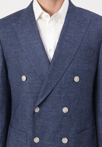 Sacou bleumarin cu două rânduri de nasturi, dintr-un material texturat, având două nasturi mari de culoare deschisă și un buzunar lateral, asortat cu o cămașă albă.
