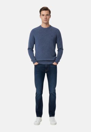 Uomo in piedi con le mani in tasca, indossa un maglione blu, jeans scuri e scarpe da ginnastica bianche su uno sfondo uniforme.