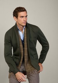 Giacca verde scuro con rever e tasca, abbinata a una sciarpa a scacchi marrone. Sotto, una camicia azzurra chiara e un gilet blu navy, pantaloni marroni strutturati con frange.