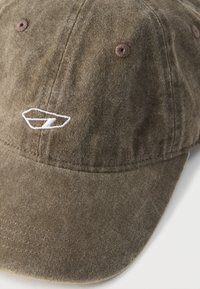 Rudas kordurovinis beisbolo kepuraitė su mažu baltu siuvinėtu logotipu priekyje ir prisiūtomis ventiliacijos skylutėmis ant skyrių.