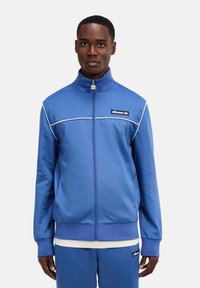 Blaues Polyester-Zip-Jacket mit hohem Kragen, kontrastierenden weißen Besätzen und einem Logo-Patch auf der Brust. Lange Ärmel mit gerippten Bündchen.