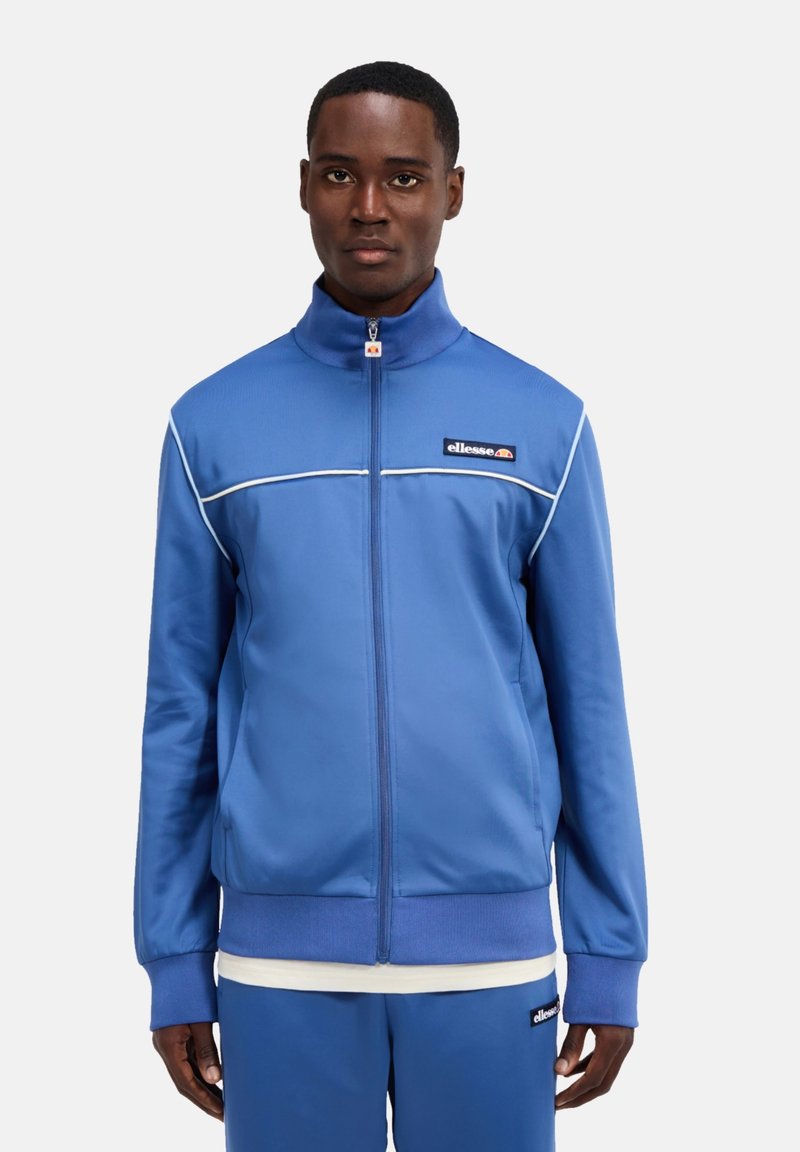 Blaues Polyester-Zip-Jacket mit hohem Kragen, kontrastierenden weißen Besätzen und einem Logo-Patch auf der Brust. Lange Ärmel mit gerippten Bündchen.