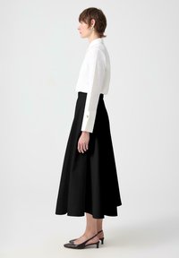 Blusa blanca de manga larga con puños abotonados, combinada con una falda negra en línea A y de largo hasta el suelo. La modelo lleva puestos unos tacones negros tipo slingback.