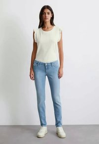 Svetlo modré rifľové nohavice s úzkym strihom a piatimi vreckami, skombinované s bielym rebrovaným pleteným topom s volánovými rukávmi. Biely tenisky dopĺňajú vzhľad.