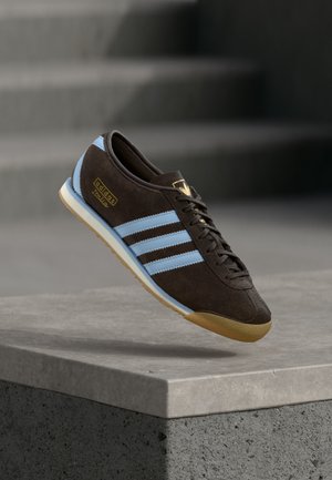 Brun Adidas sneaker med lyseblå striber og gummisål, hængende over en betonoverkant med slørede trapper i baggrunden.