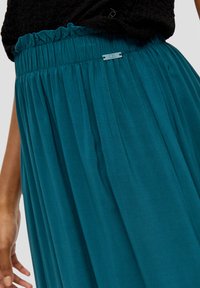 Teal, geplooide maxi rok met een elastische tailleband, met een subtiele glans en een klein metalen label met de tekst "QS" op de taille.